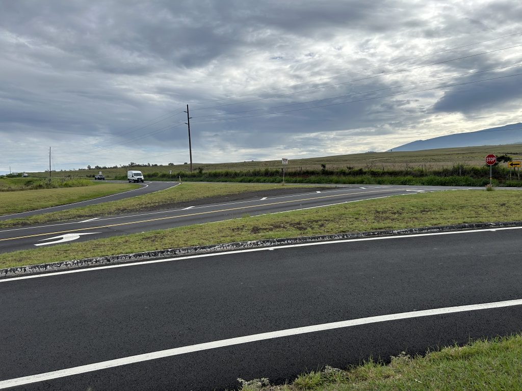 Turning lanes onto road with grass in the foreground and background and cloudy sky.